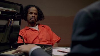 Movie still from “The Wire” (2002), created by David Simon – A man in a red shirt sitting in front of a judge; Medium shot, Over the shoulder angle
