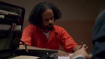 Movie still from “The Wire” (2002), created by David Simon – A man in a red prison suit writing on a paper; Medium shot, High angle