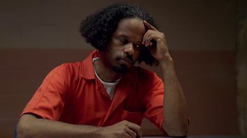 Movie still from “The Wire” (2002), created by David Simon – A man in red shirt sitting at a table with his head resting on his hand; Close Up shot, High angle