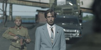 Movie still from “Chernobyl” (2019), directed by Johan Renck – A man in a suit and tie standing in front of a car; Medium shot, Over the shoulder angle