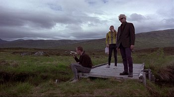 Movie still from “Trainspotting” (1996), directed by Danny Boyle – A man sitting on a wooden platform next to two other people; Wide shot, Low angle