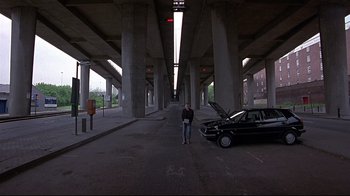 Movie still from “Trainspotting” (1996), directed by Danny Boyle – A man standing next to a car in an empty parking lot; Wide shot, Low angle