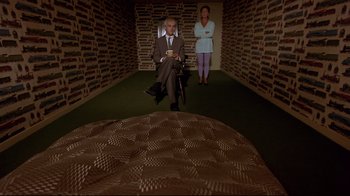 Movie still from “Trainspotting” (1996), directed by Danny Boyle – A man sitting on a chair in a room with a woman standing in the background; Wide shot, High angle