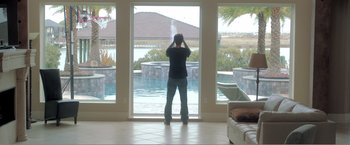 Movie still from “99 Homes” (2014), directed by Ramin Bahrani – A man standing in front of an open window looking out at a pool; Wide shot, Low angle