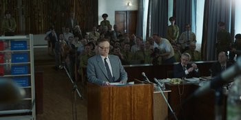 Movie still from “Chernobyl” (2019), directed by Johan Renck – A man in a gray suit is speaking in front of a group of people; Medium shot, Over the shoulder angle