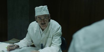 Movie still from “Chernobyl” (2019), directed by Johan Renck – A man in a white lab coat sitting in front of a table; Medium shot, Over the shoulder angle