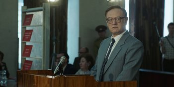 Movie still from “Chernobyl” (2019), directed by Johan Renck – A man in a gray suit and tie is speaking; Medium shot, Over the shoulder angle