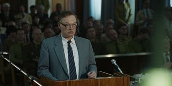 Movie still from “Chernobyl” (2019), directed by Johan Renck – A man in a gray suit and tie is speaking at a podium; Medium shot, Over the shoulder angle