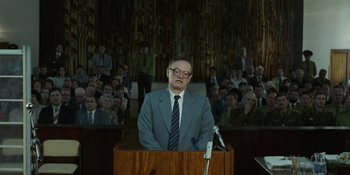 Movie still from “Chernobyl” (2019), directed by Johan Renck – A man in a suit and tie standing at a podium in front of an audience; Medium shot, Low angle