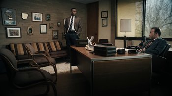 Movie still from “True Detective” (2014), created by Issa López – A man in a suit and tie standing in front of a desk in an office; Wide shot, High angle