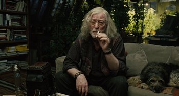 Movie still from “Children of Men” (2006), directed by Alfonso Cuarón – An older man smoking a cigarette while sitting on a couch; Medium shot, Low angle