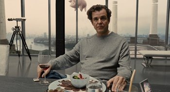 Movie still from “Children of Men” (2006), directed by Alfonso Cuarón – A man sitting at a table with a plate of food and a glass of wine in front of him; Medium shot, Over the shoulder angle