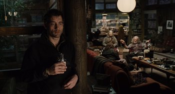 Movie still from “Children of Men” (2006), directed by Alfonso Cuarón – A man holding a drink while standing next to a wall; Medium shot, Over the shoulder angle