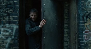 Movie still from “Children of Men” (2006), directed by Alfonso Cuarón – A man standing in a doorway smiling for the camera; Medium shot, Low angle