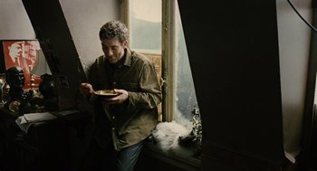 Movie still from “Children of Men” (2006), directed by Alfonso Cuarón – A man standing in front of a window with a plate of food; Medium shot, Low angle