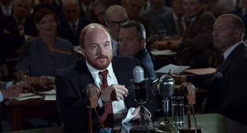 Movie still from “Trumbo” (2015), directed by Jay Roach – A man sitting at a table with microphones; Medium shot, Over the shoulder angle