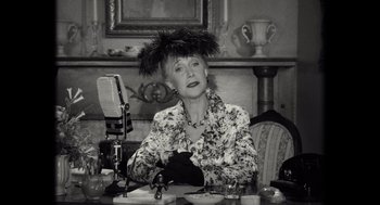 Movie still from “Trumbo” (2015), directed by Jay Roach – An older woman sitting at a table in front of a microphone; Medium shot, Low angle