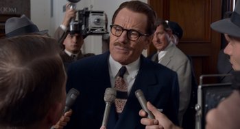 Movie still from “Trumbo” (2015), directed by Jay Roach – A man in a suit and tie talking into microphones in front of other men; Close Up shot, Over the shoulder angle