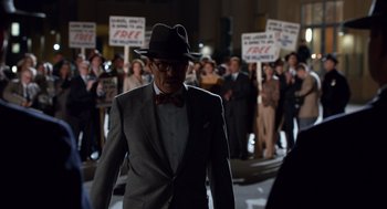 Movie still from “Trumbo” (2015), directed by Jay Roach – A man in a suit and a hat walking in front of a group of people; Wide shot, High angle