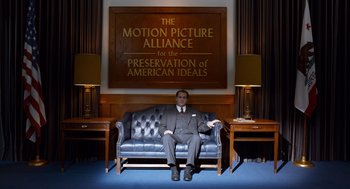 Movie still from “Trumbo” (2015), directed by Jay Roach – A man sitting on a blue couch in front of an american ideals sign; Wide shot, High angle