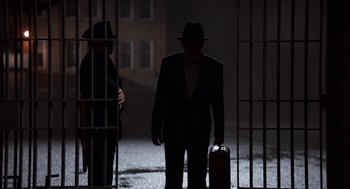Movie still from “Trumbo” (2015), directed by Jay Roach – A man in a suit and a hat is standing next to a man in a jail cell; Wide shot, Low angle