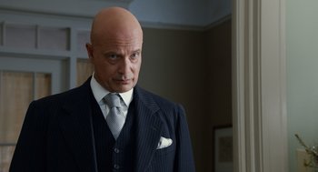 Movie still from “Trumbo” (2015), directed by Jay Roach – A bald man in a suit and tie looking at the camera; Close Up shot, Low angle