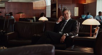Movie still from “Up in the Air” (2009), directed by Jason Reitman – A man in a suit and tie sitting on a couch talking on a cell phone; Medium shot, Over the shoulder angle