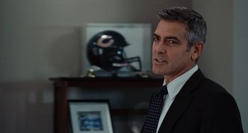 Movie still from “Up in the Air” (2009), directed by Jason Reitman – A man in a suit and tie in front of a football helmet; Close Up shot, Over the shoulder angle