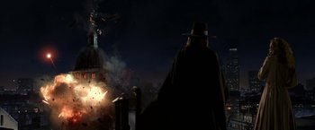 Movie still from “V for Vendetta” (2005), directed by James McTeigue – A man wearing a hat and a long black coat standing in front of fireworks; Extreme Wide shot, Low angle