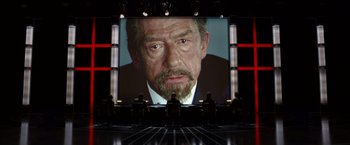 Movie still from “V for Vendetta” (2005), directed by James McTeigue – People sitting in front of a large screen with a picture of an older man; Medium shot, Over the shoulder angle