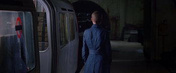 Movie still from “V for Vendetta” (2005), directed by James McTeigue – A man in a blue coat standing in front of a train; Medium shot, Over the shoulder angle
