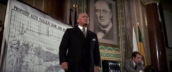 Movie still from “Chinatown” (1974), directed by Roman Polanski – An older man wearing a suit and tie in front of a large portrait of a man; Medium shot, Low angle