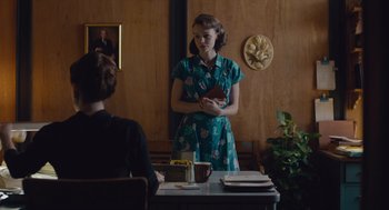 Movie still from “Wildlife” (2018), directed by Paul Dano – A woman standing in front of a table holding a book; Medium shot, Over the shoulder angle