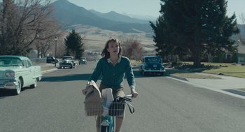 Movie still from “Wildlife” (2018), directed by Paul Dano – A woman riding a bike down a street with mountains in the background; Wide shot, Over the shoulder angle