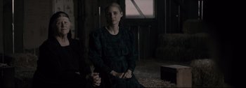 Movie still from “Women Talking” (2022), directed by Sarah Polley – A woman in a black dress sitting in a barn; Medium shot, Over the shoulder angle