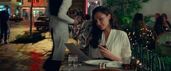 Movie still from “Wonder Woman 1984” (2020), directed by Patty Jenkins – A woman sitting at a table holding a glass of wine; Medium shot, Over the shoulder angle