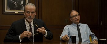 Movie still from “Zodiac” (2007), directed by David Fincher – Two men are sitting at a table and one of the men is holding a pair of glasses; Medium shot, Over the shoulder angle