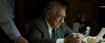 Movie still from “Zodiac” (2007), directed by David Fincher – An older man in a suit and tie looking at a piece of paper; Medium shot, High angle