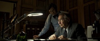 Movie still from “Zodiac” (2007), directed by David Fincher – A man standing next to another man at a desk; Medium shot, Low angle