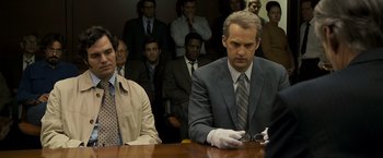 Movie still from “Zodiac” (2007), directed by David Fincher – A man sitting at a table in front of a group of people; Medium shot, Over the shoulder angle