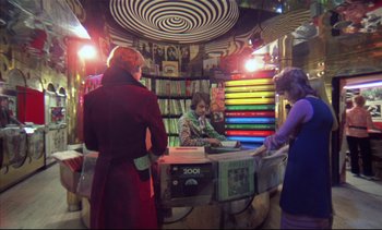 Movie still from “A Clockwork Orange” (1971), directed by Stanley Kubrick – Three people standing in front of a table with books on top of it; Wide shot, High angle