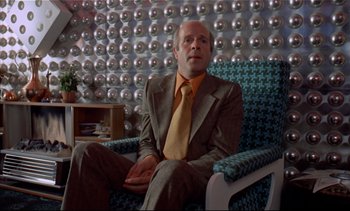 Movie still from “A Clockwork Orange” (1971), directed by Stanley Kubrick – A man in a suit and tie sitting in a chair; Medium shot, Low angle