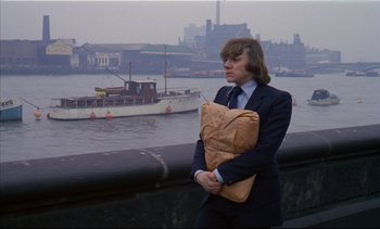 Movie still from “A Clockwork Orange” (1971), directed by Stanley Kubrick – A man in a suit holding a paper bag in front of a body of water; Medium shot, Low angle