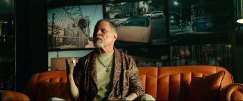 Movie still from “Ambulance” (2022), directed by Michael Bay – An older man sitting on a couch in front of a tv; Medium shot, Low angle