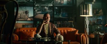 Movie still from “Ambulance” (2022), directed by Michael Bay – A man sitting on top of an orange couch; Medium shot, Low angle