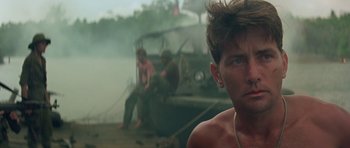 Movie still from “Apocalypse Now” (1979), directed by Francis Ford Coppola – A shirtless man sitting next to a boat in the water; Close Up shot, Low angle