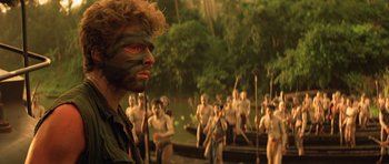 Movie still from “Apocalypse Now” (1979), directed by Francis Ford Coppola – A man with a painted face is standing in front of a group of people; Close Up shot, Over the shoulder angle