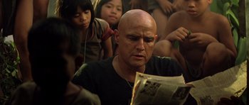 Movie still from “Apocalypse Now” (1979), directed by Francis Ford Coppola – A man with bald head reading a newspaper while sitting in a crowd of people; Close Up shot, Low angle