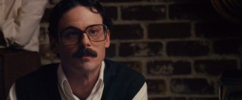 Movie still from “Argo” (2012), directed by Ben Affleck – A man wearing glasses and a sweater sitting in front of a brick wall; Close Up shot, Low angle