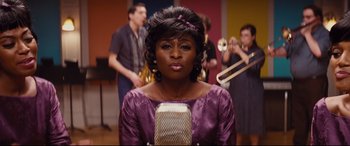 Movie still from “Bad Times at the El Royale” (2018), directed by Drew Goddard – A woman in a purple dress is holding a microphone; Close Up shot, Over the shoulder angle